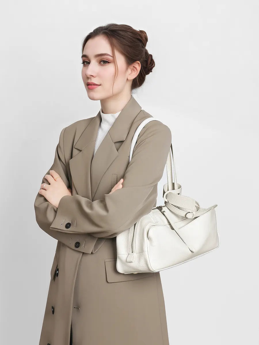 Woman in a beige trench coat holding a white handbag against a plain background