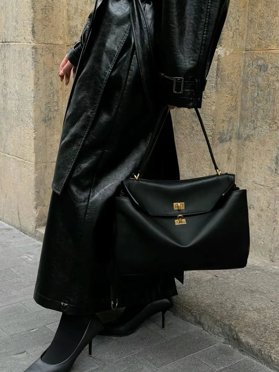 Person wearing a black coat and holding a black handbag against a stone wall.