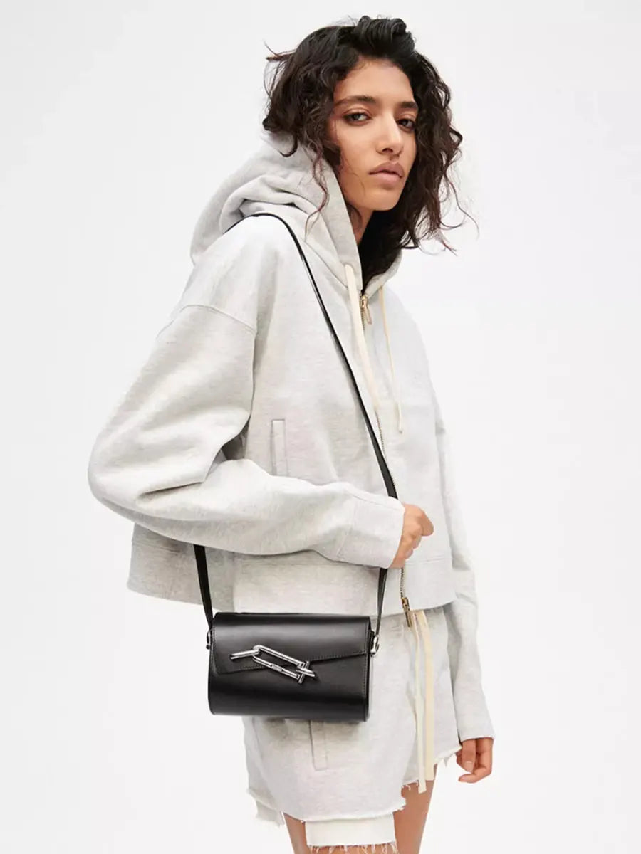 Woman wearing a light gray hoodie and white pants, holding a black handbag with a silver clasp on a white background.