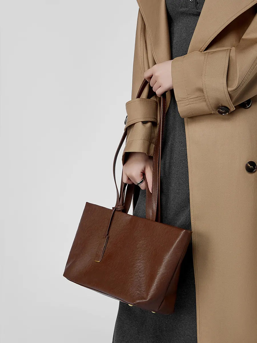 Person wearing a brown coat holding a brown leather handbag against a plain background