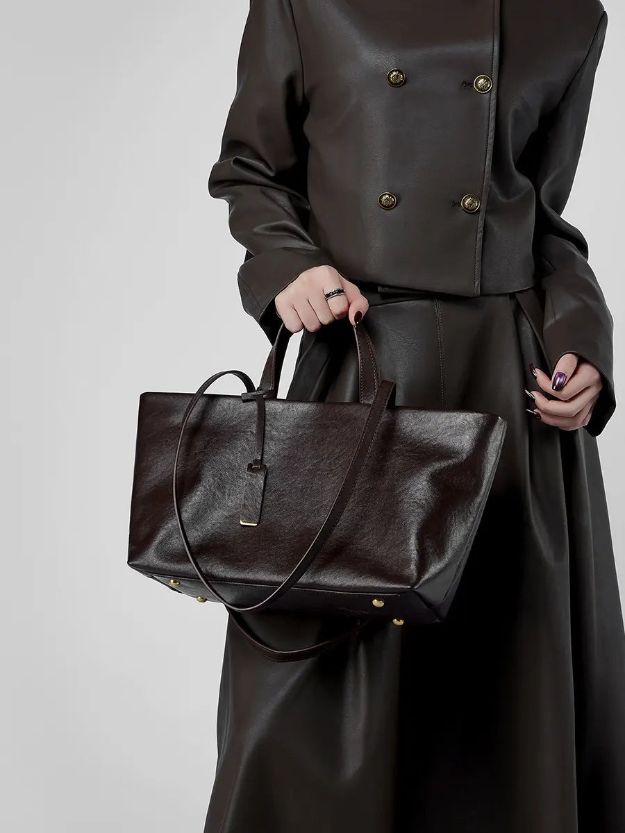 Person wearing a dark coat holding a matching dark handbag against a neutral background