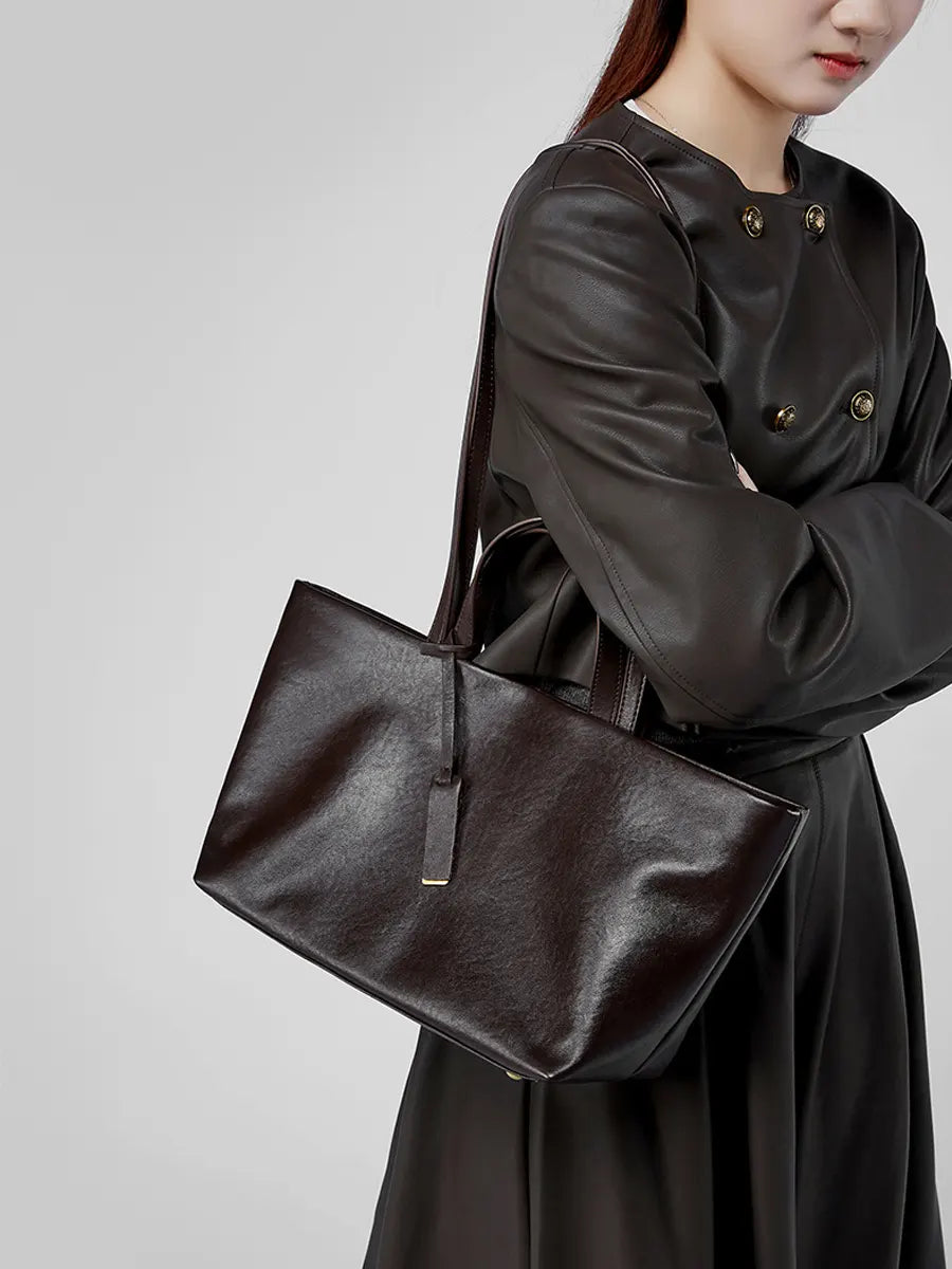 Person wearing a dark coat holding a matching dark handbag against a plain background