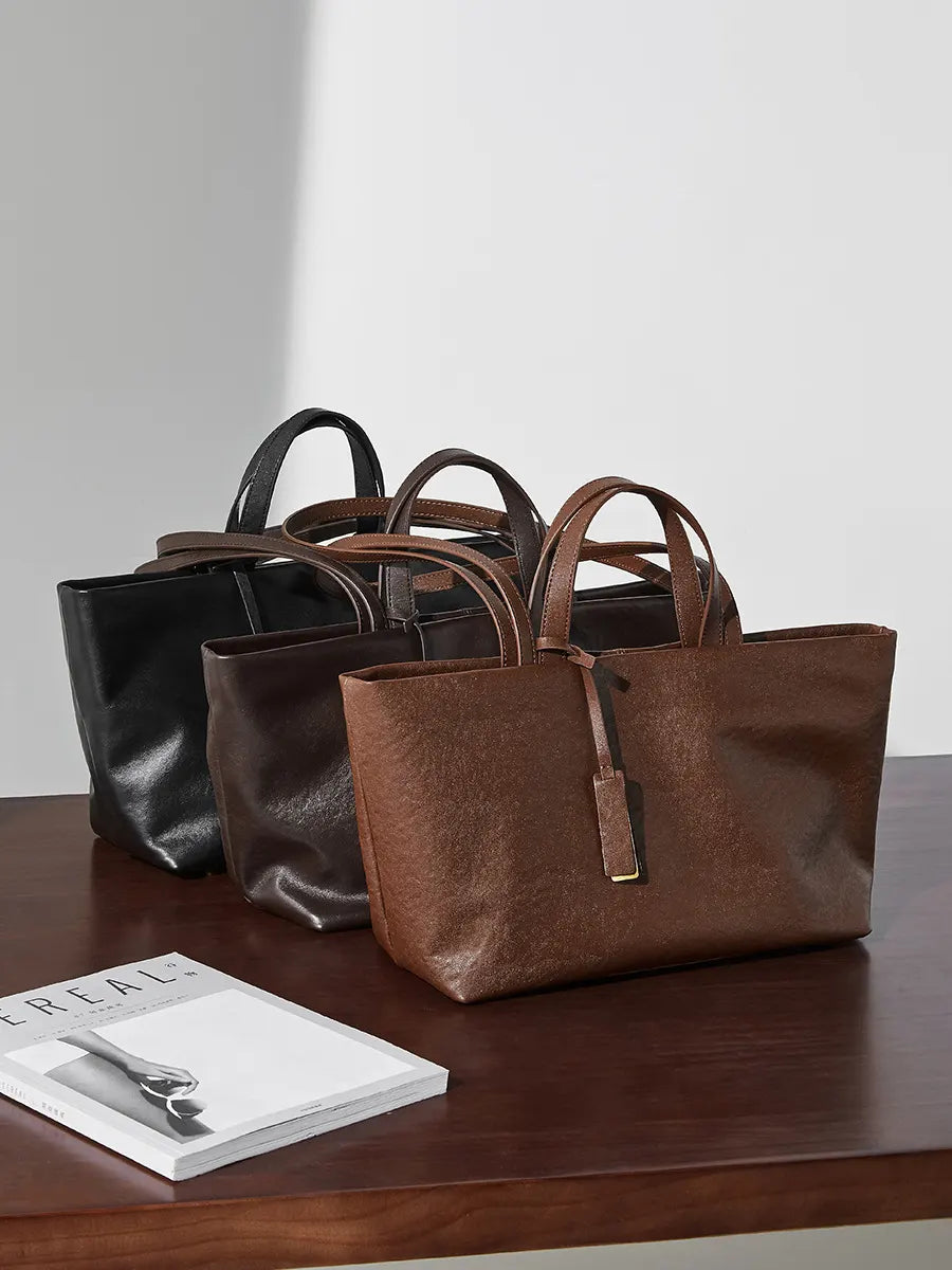 Three leather handbags in black, brown, and another brown shade on a wooden surface with a magazine underneath.