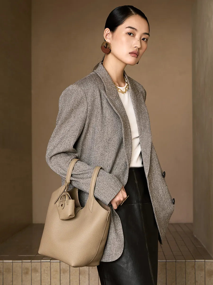 Woman holding a beige handbag wearing a gray coat against a neutral background