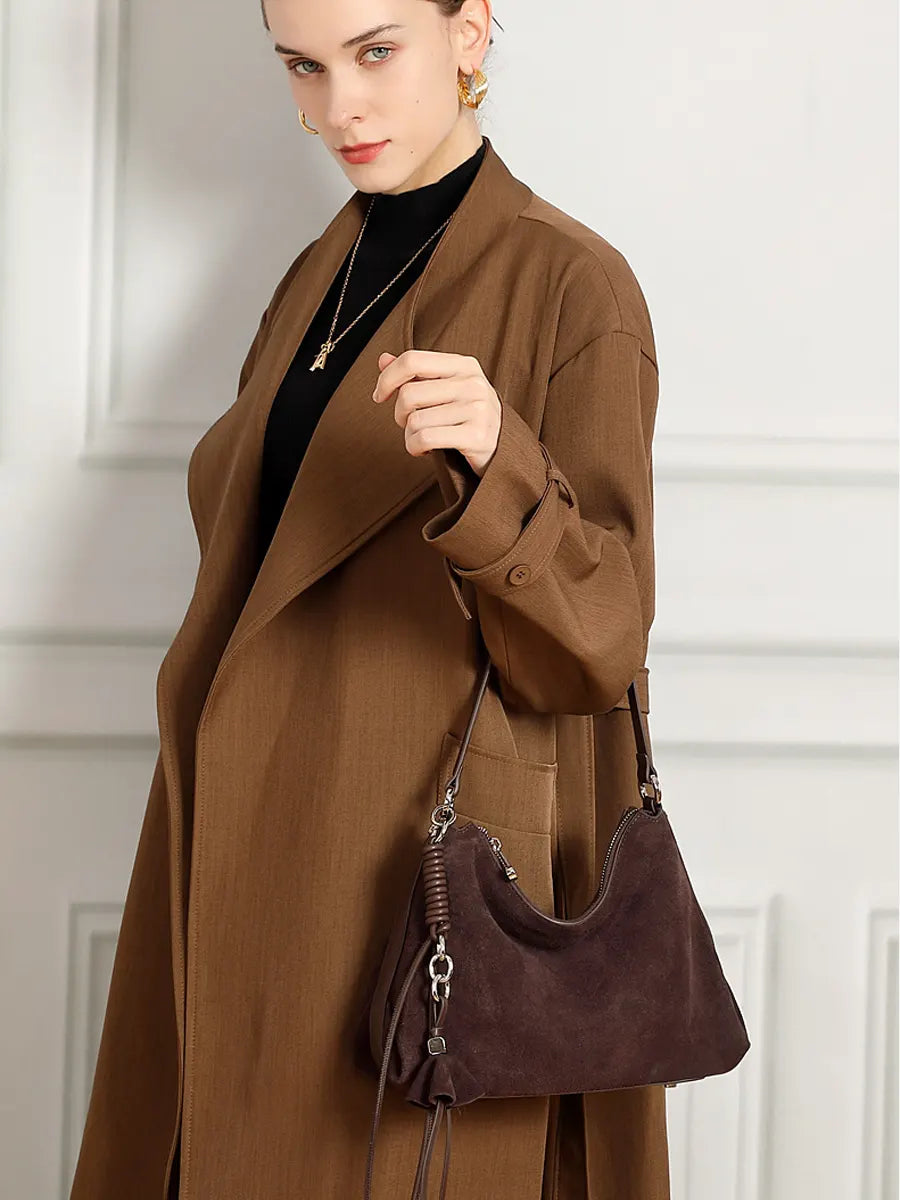 Woman in a brown coat holding a brown handbag against a white wall